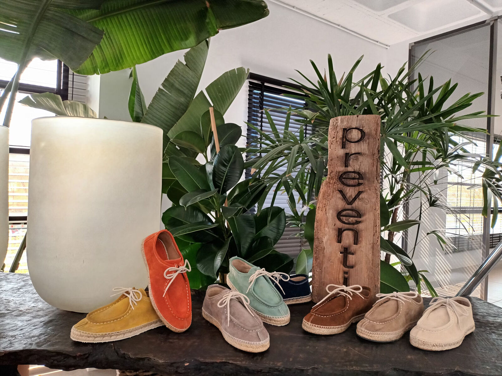 Zapatos de ante en varios colores con suela de yute, junto a un cartel de madera con la palabra Preventi.