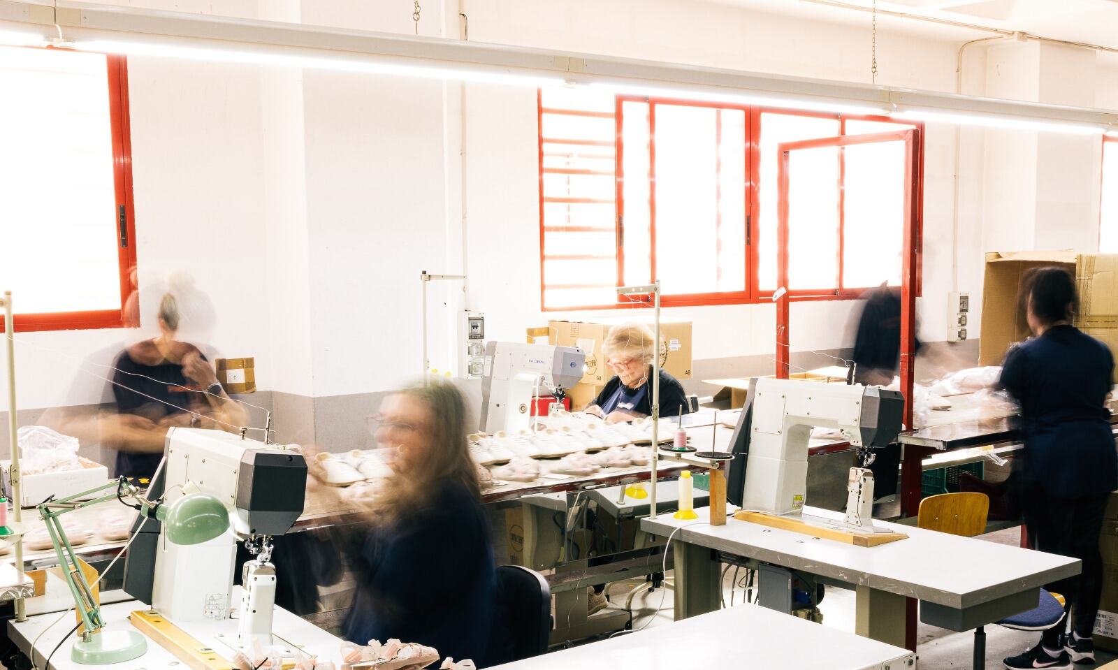 Sala de costura con máquinas de columna y operarias trabajando frente a ventanas de marco rojo.
